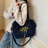 A woman wearing a light gray sweatshirt and blue jeans holds a dark denim crossbody bag with gold chain details.