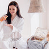 A woman sits on a bed with a white robe and a multi-compartment travel organizer bag open beside her.