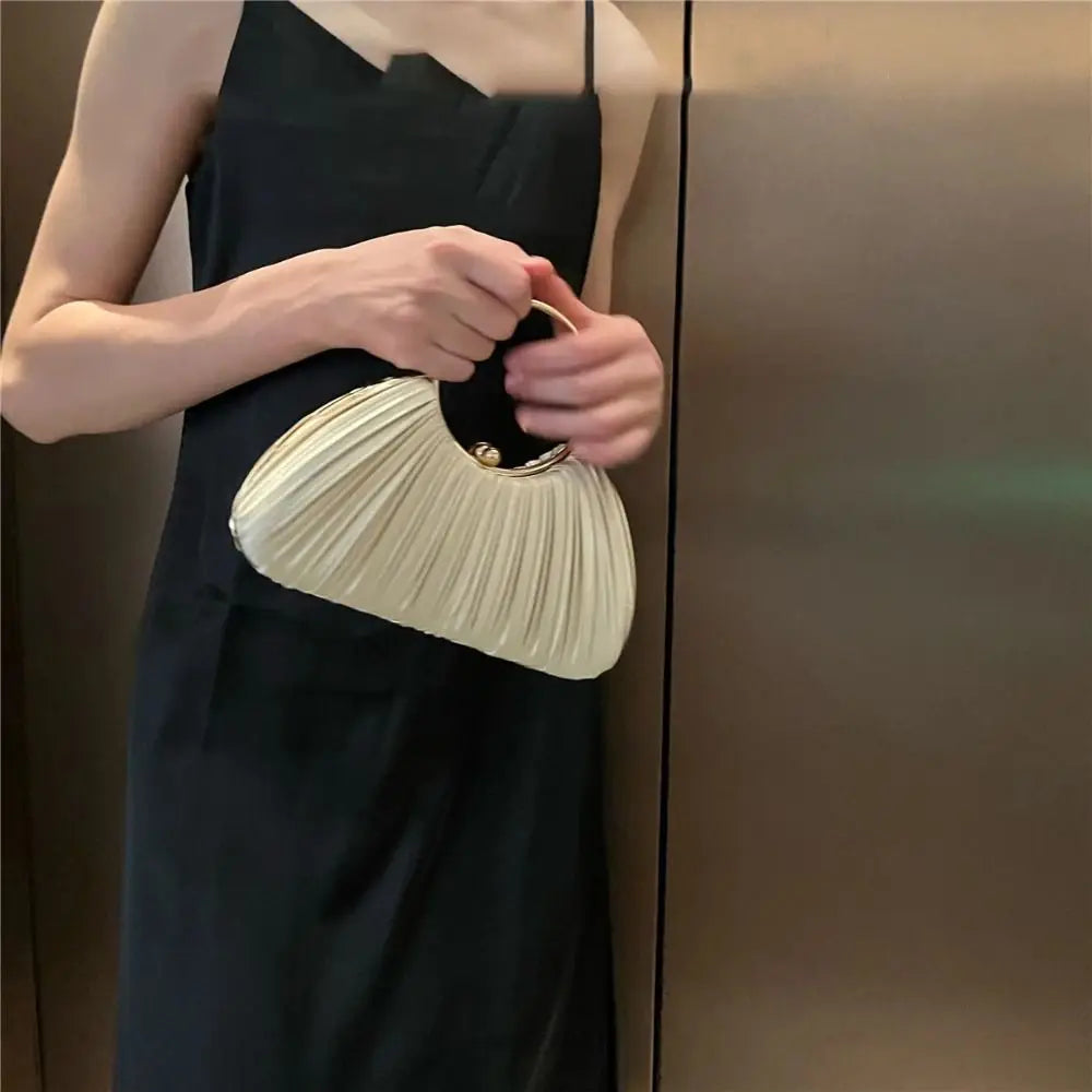 A woman holds an elegant satin pleated evening clutch bag in gold against a dark dress.