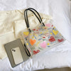 A clear plastic tote bag with a black strap and heart-shaped confetti decoration is shown next to a smartphone.