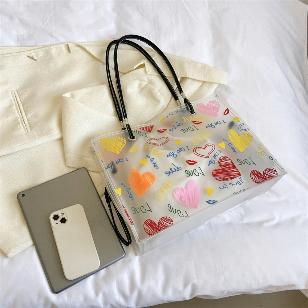 A clear plastic tote bag with a black strap and heart-shaped confetti decoration is shown next to a smartphone.