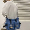 A light blue nylon crossbody bag with black accents is shown from the back, hanging against a white window blind.
