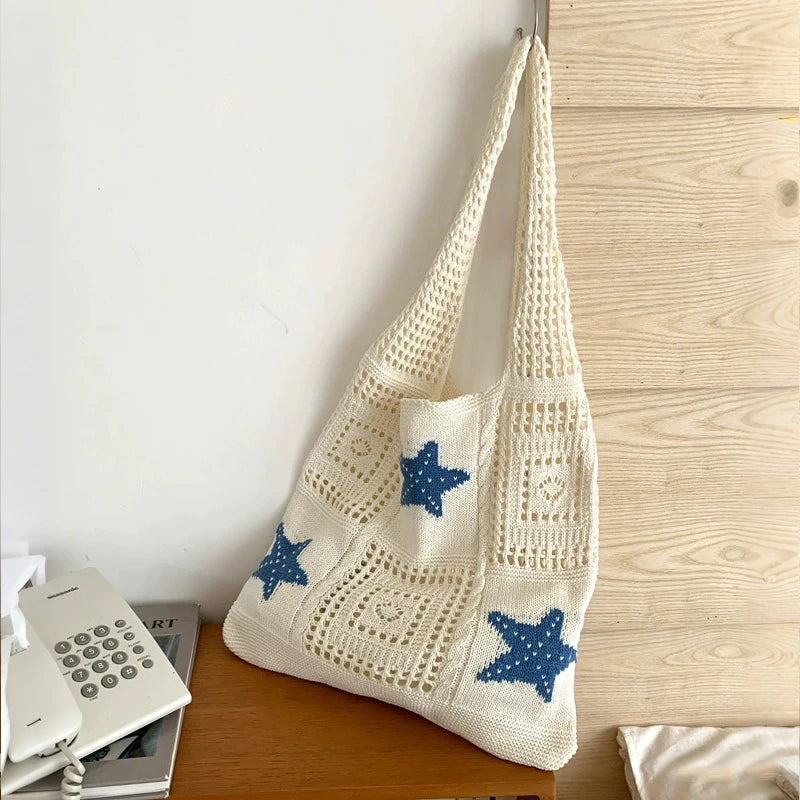 A cream-colored knitted tote bag with blue star patterns hanging next to a wooden surface.