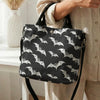 A woman holds a black tote bag with a white bat print and a black strap against a light-colored shirt.