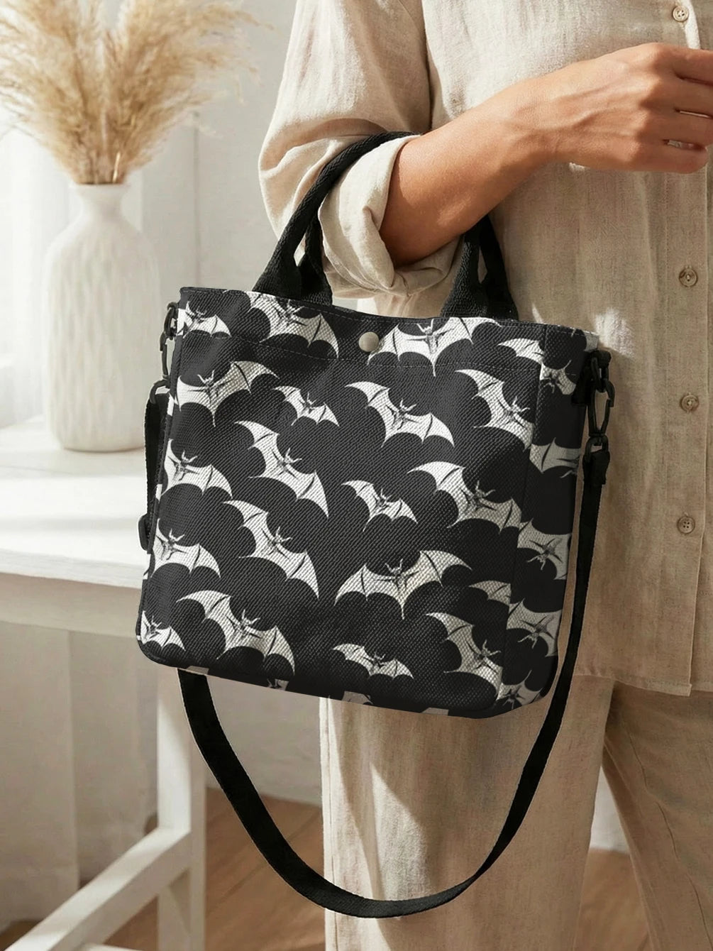 A woman holds a black tote bag with a white bat print and a black strap against a light-colored shirt.