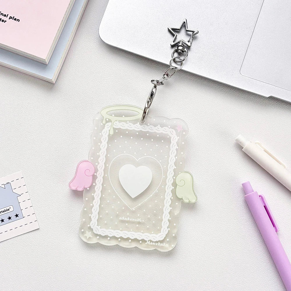 A clear acrylic photocard holder with a heart design and angel wings is attached to a silver charm.