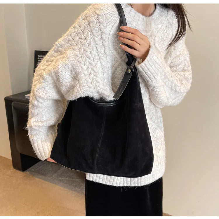 A woman wearing a cream knit sweater and black skirt holds a black suede slouchy shoulder bag.