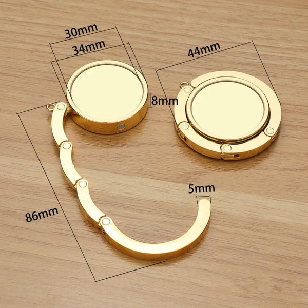 A gold-colored folding alloy handbag hanger hook with various dimensions shown on a wooden surface.