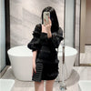 A woman wearing a black ruffled dress and holding a black chain shoulder clutch bag in a modern bathroom setting.