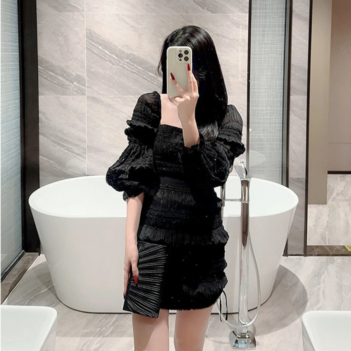 A woman wearing a black ruffled dress and holding a black chain shoulder clutch bag in a modern bathroom setting.