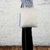Un sac fourre-tout en coton blanc est exposé accroché à un vêtement contre un mur de briques blanches.