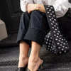 A woman wearing a white shirt and dark jeans holds a black leather shoulder bag adorned with silver metal rivets.