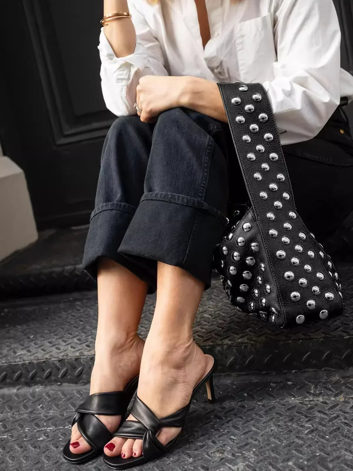 A woman wearing a white shirt and dark jeans holds a black leather shoulder bag adorned with silver metal rivets.