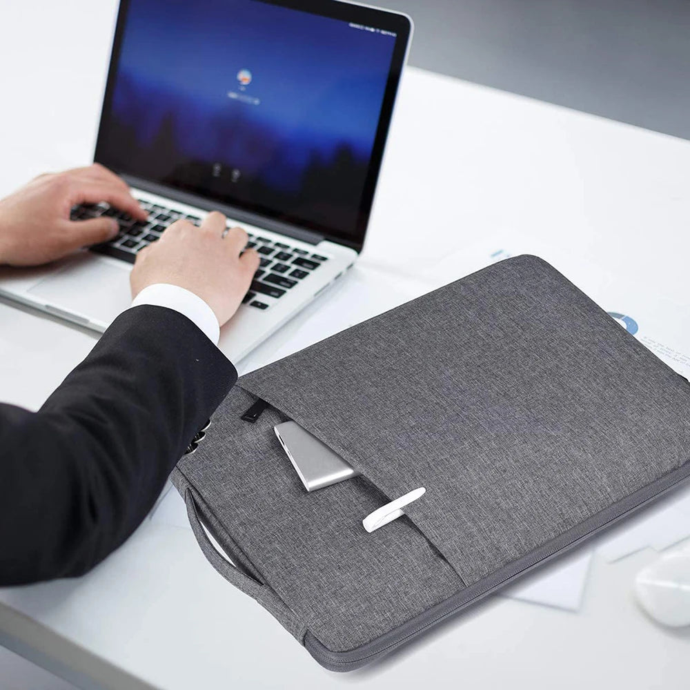 A person places a gray laptop sleeve containing a silver item on a white desk next to an open laptop.