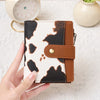 A hand holds a women's RFID-blocking compact bifold zipper wallet with a black and white cow print design.