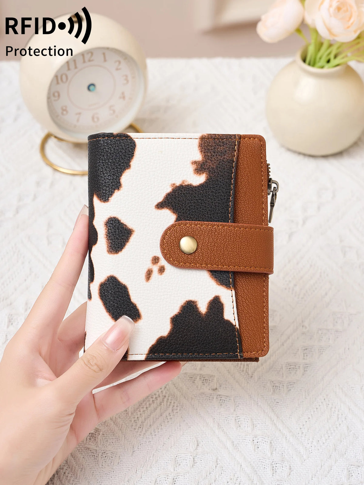 A hand holds a women's RFID-blocking compact bifold zipper wallet with a black and white cow print design.