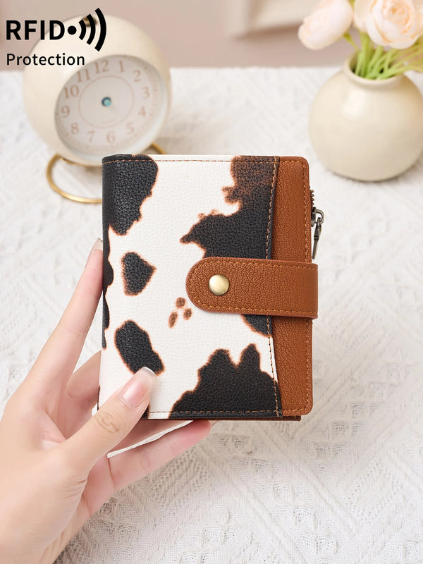 A hand holds a women's RFID-blocking compact bifold zipper wallet with a black and white cow print design.