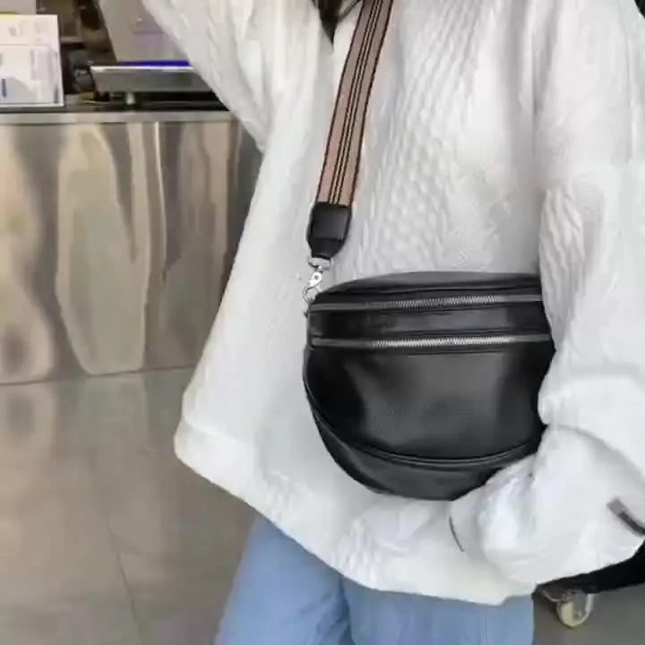 A person is holding a black soft leather crossbody bag with double zippers and an adjustable strap against a white jacket.