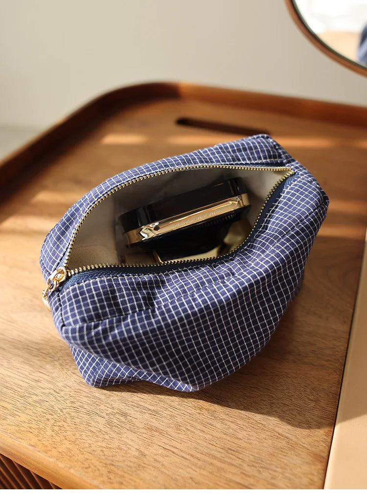 Pochette en tissu à carreaux bleu marine avec fermeture éclair dorée, contenant un produit cosmétique noir, posé sur une surf
