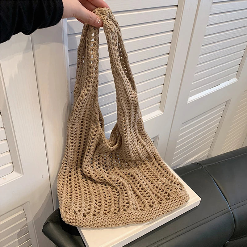 A beige bohemian knit shoulder bag is being held by a hand, resting on a white surface.