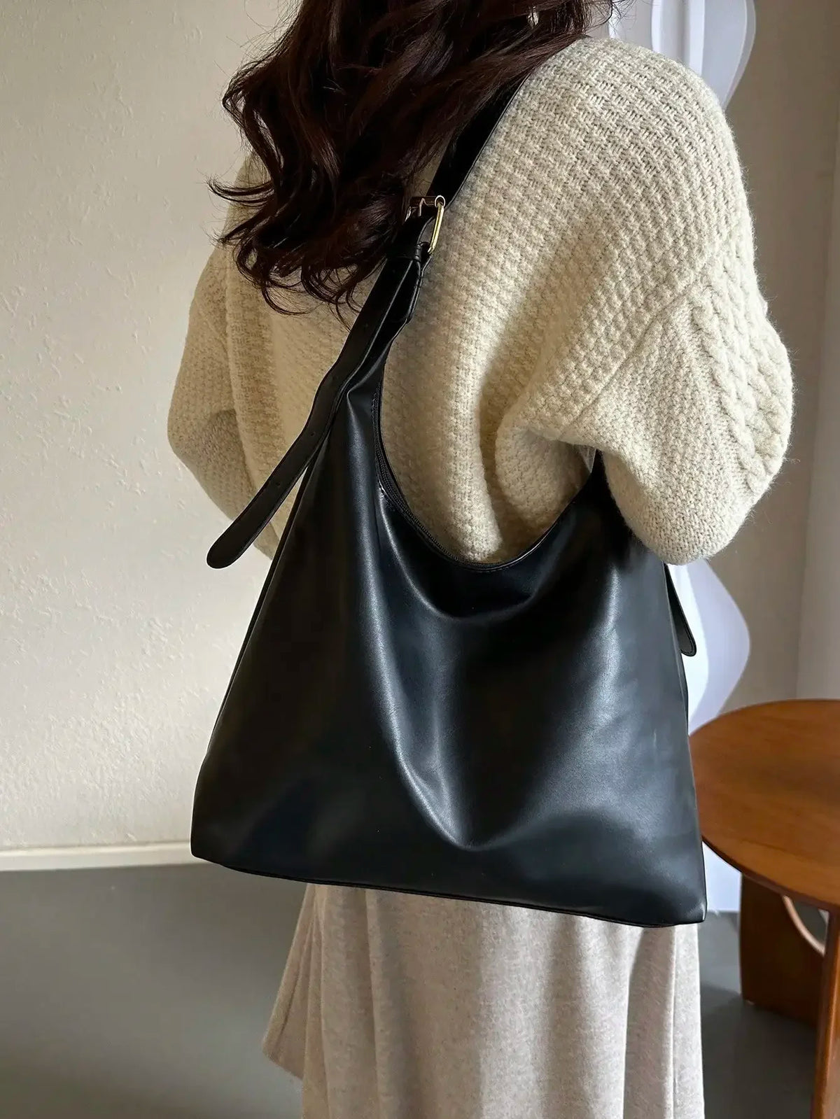 A black PU leather slouchy hobo shoulder bag carried by a person wearing a cream knit sweater.