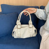 A hand is holding a white PU leather shoulder bag with silver rivets on a blue sofa.