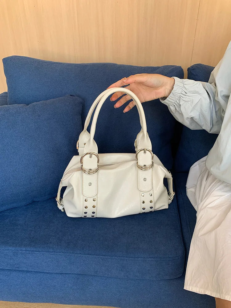 A hand is holding a white PU leather shoulder bag with silver rivets on a blue sofa.