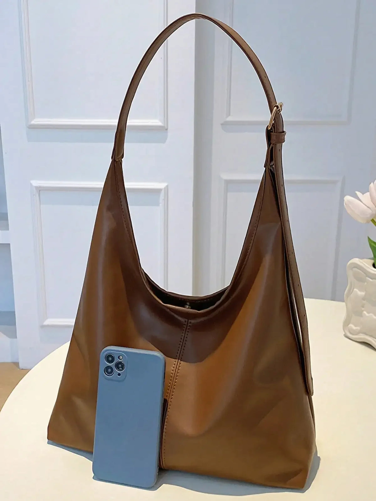 A brown PU leather slouchy hobo shoulder bag resting on a white surface next to a smartphone.
