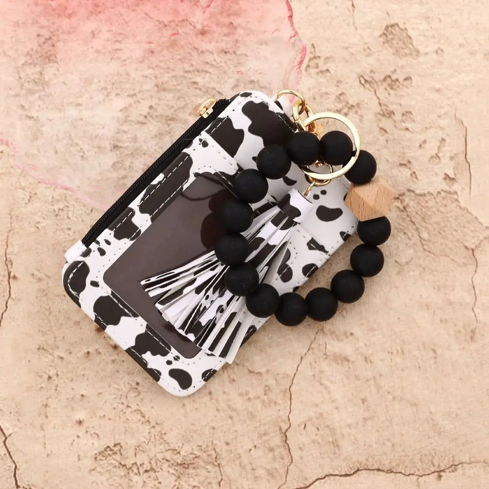 A black and white cow print card holder wallet with a gold tassel keychain and black beads on a light background.