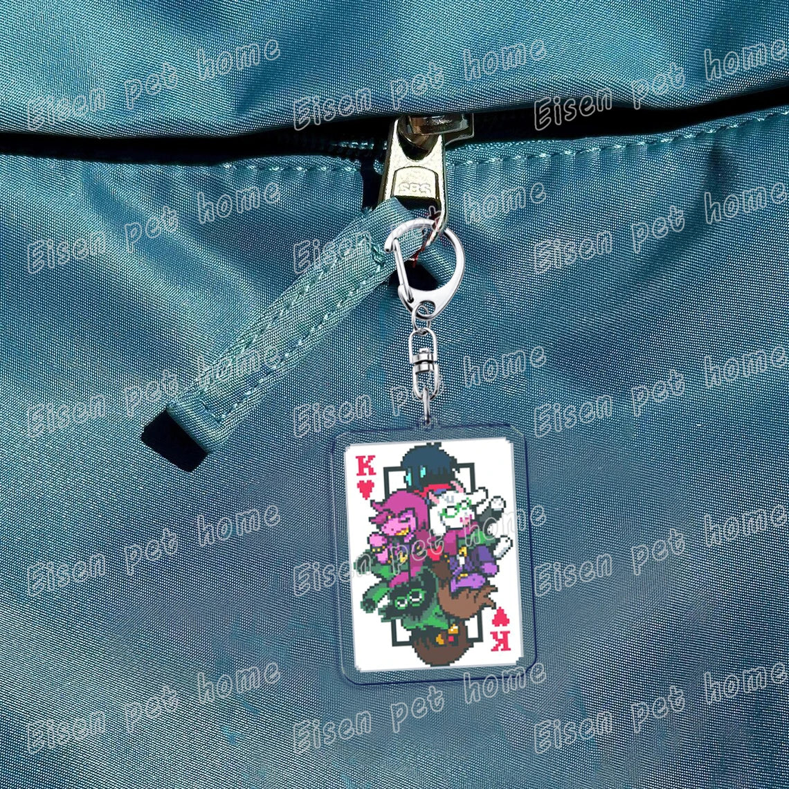 A keychain featuring Deltarune chibi poker cards with a K symbol on a light blue fabric background.
