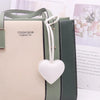 A light beige leather pouch with a white heart charm and dark green leather straps.