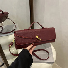 A burgundy crocodile-embossed PU leather clutch bag with a gold-toned strap is displayed on a white surface.