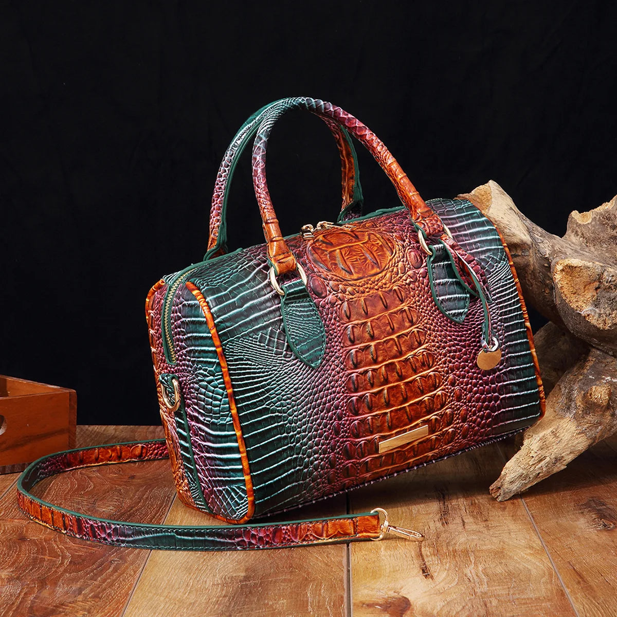 A luxury crocodile pattern crossbody tote bag in green and brown leather resting on a wooden surface next to a piece of drift