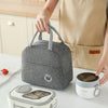 A person holds a gray insulated lunch bag next to a ceramic mug and a white food container on a table.