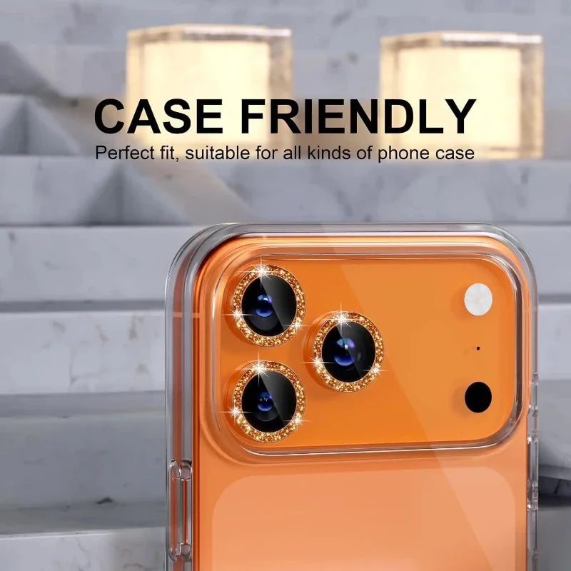 An orange iPhone with a glitter diamond camera lens protector and metal frame, shown against a marble background.