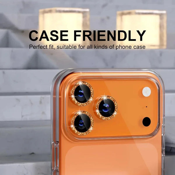 An orange iPhone with a glitter diamond camera lens protector and metal frame, shown against a marble background.