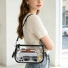 Une femme tient un sac à l'épaule en PVC transparent avec une sangle en cuir marron et un smartphone dedans.