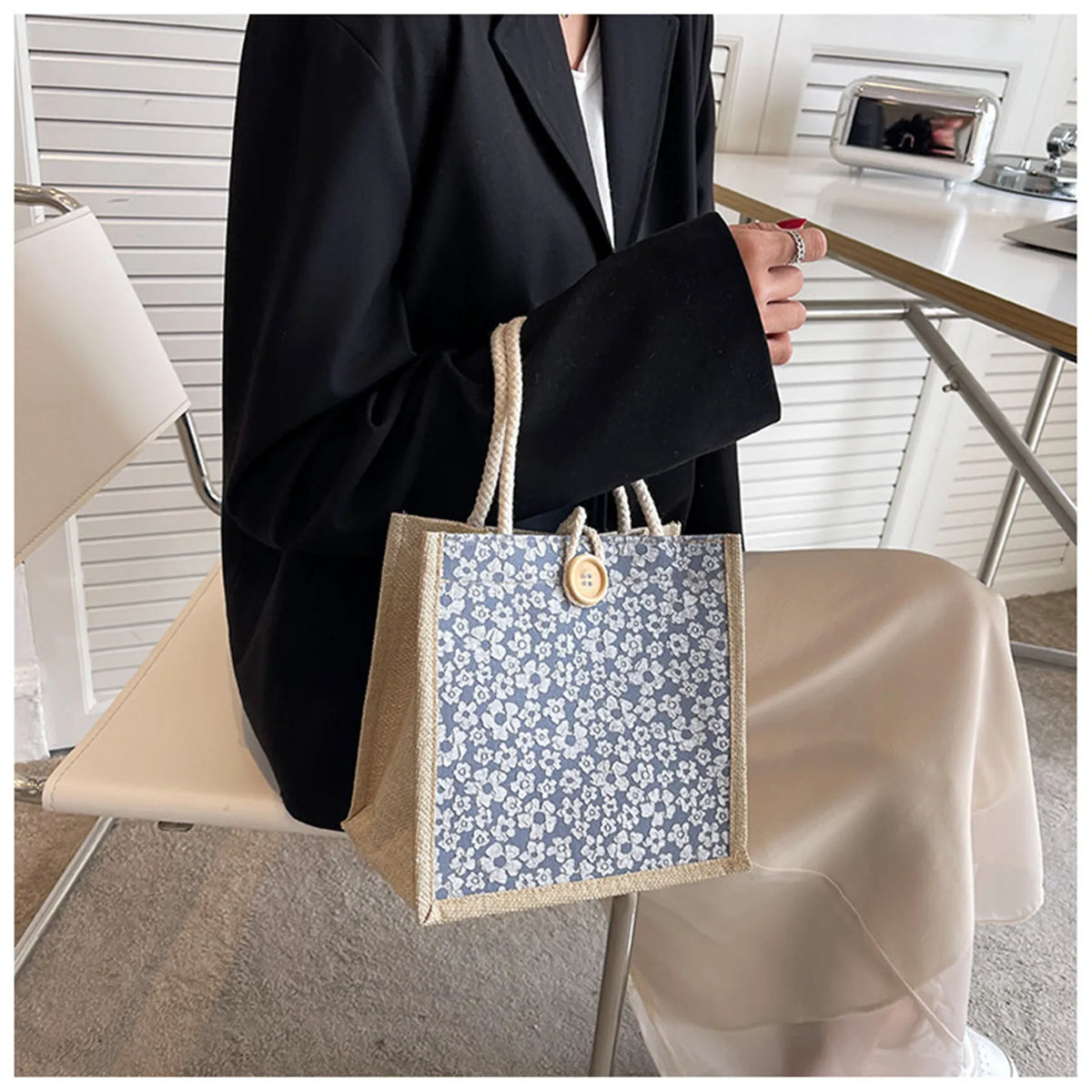 A beige floral linen tote bag with blue accents is held by a person in a black blazer.