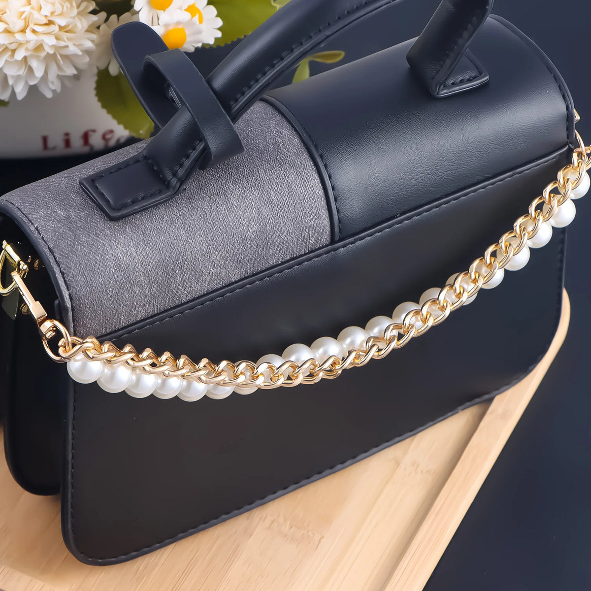 A dark gray and black leather shoulder bag with a gold pearl chain strap resting on a wooden surface.