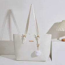 A cream-colored PU leather shoulder tote bag with light beige straps and a fluffy pom pom charm.