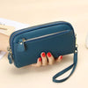 A dark teal leather double-zip wristlet wallet with a gold-toned strap is held by a hand on a light background.