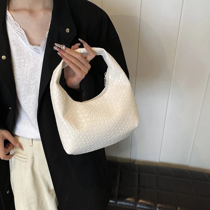 A person holds a cream woven embossed hobo shoulder bag with a top handle against a white background.
