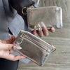 A hand holds a silver metallic leather compact coin purse and key holder against a gray suit jacket.