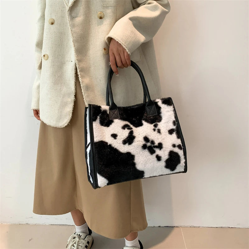 A woman holds a black and white leopard print faux fur designer shoulder tote bag while wearing a beige coat and skirt.