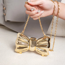 A hand holds a gold heart-shaped acrylic evening clutch with a chain strap on a light surface.