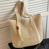 A natural straw woven tote bag hanging against a white slatted background.