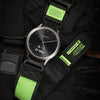 A black Garmin watch with a green silicone strap and a green protective cover is shown on a black fabric background.