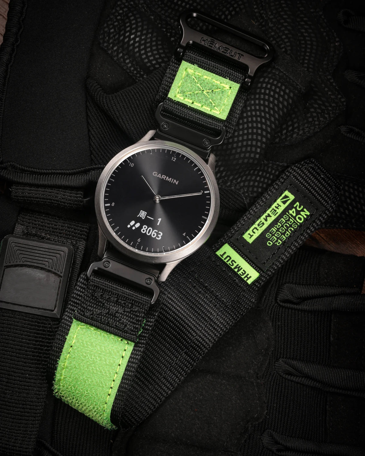 A black Garmin watch with a green silicone strap and a green protective cover is shown on a black fabric background.