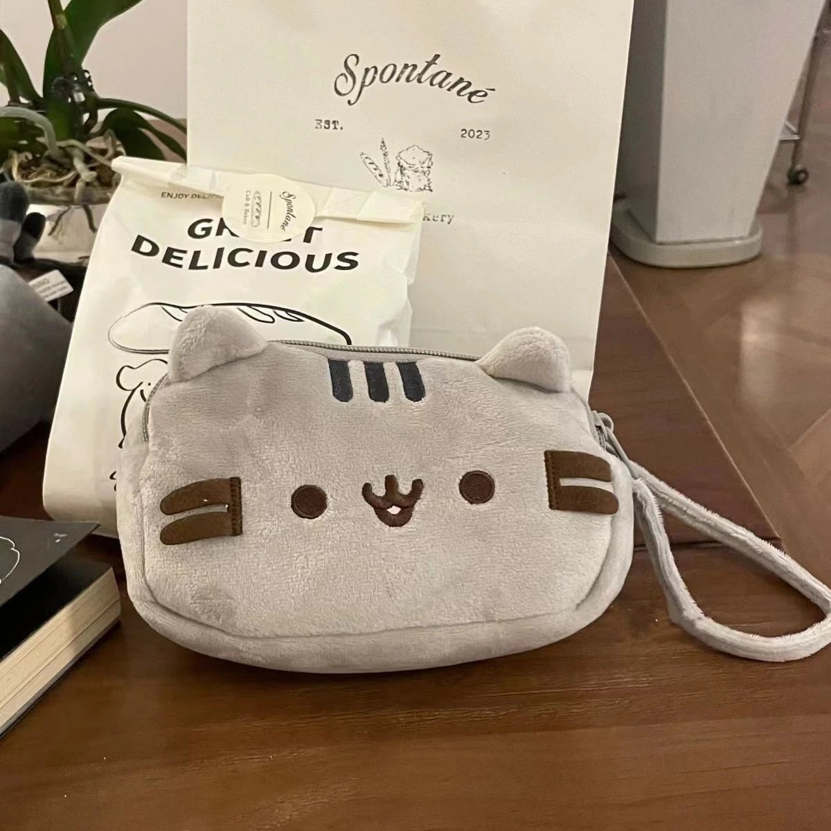 A soft, light gray cat-shaped storage pouch with brown accents is displayed on a wooden surface.