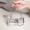 A hand holds a silver acrylic bow clasp mini handbag with a chain strap on a light surface.